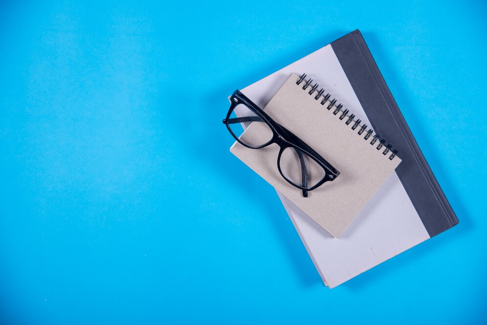 Fundo azul com dois cadernos beges e um óculos preto empilhados no centro mais à direita, que é por onde começar a estudar para o ENEM